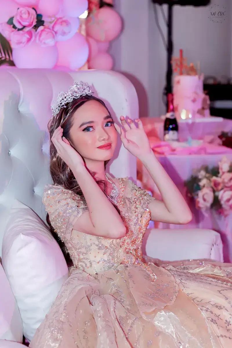 Debutante seated in a full ball gown, touching her hair, against white and pink decor