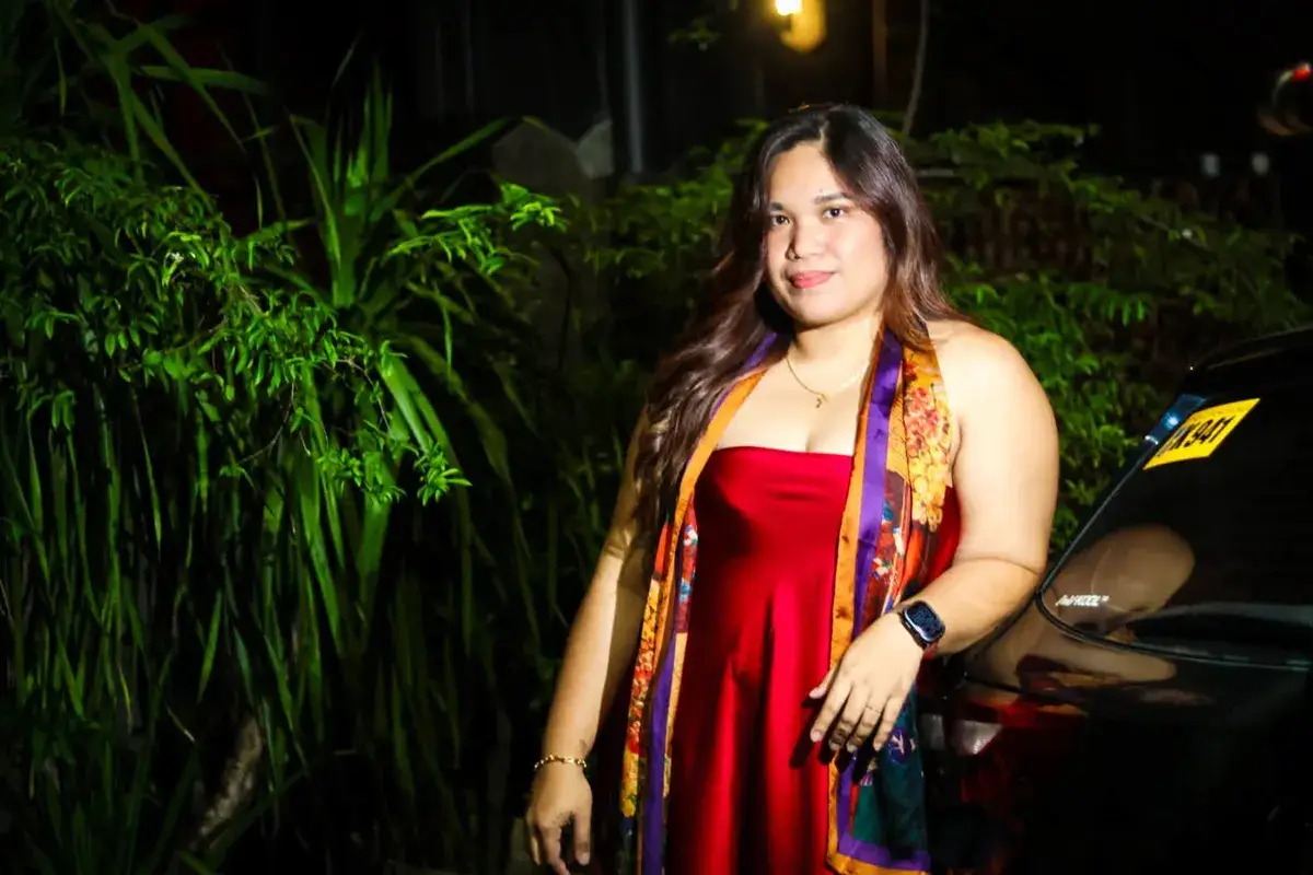 Woman in a red dress and colourful scarf among plants, lit at night